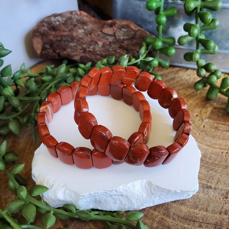 K&ouml;nnte beinhalten: Zwei Armb&auml;nder aus rotem Jaspis. Jedes Armband besteht aus rechteckigen, polierten Steinen. Die Armb&auml;nder werden auf einer wei&szlig;en, strukturierten Oberfl&auml;che mit gr&uuml;nem Laub und einem Holzst&uuml;ck im Hintergrund pr&auml;sentiert.