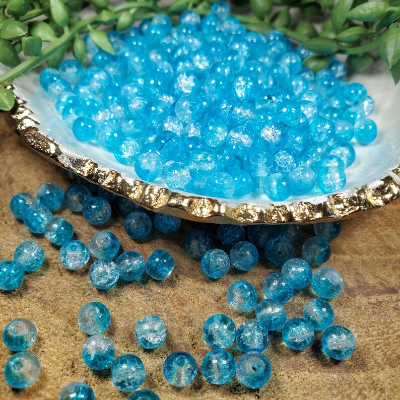 Pode incluir: Uma cole&ccedil;&atilde;o de contas de vidro azul claro com acabamento craquelado. As contas est&atilde;o espalhadas sobre uma superf&iacute;cie de madeira e em um prato decorativo em forma de concha com borda dourada. Folhagem verde &eacute; vis&iacute;vel no fundo.