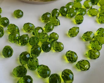 Puede incluir: Una colección de pequeñas cuentas de vidrio de color verde oliva facetadas esparcidas sobre una superficie. Cada cuenta presenta una superficie con múltiples facetas que refleja la luz, creando un aspecto brillante. Las cuentas son translúcidas y uniformes.