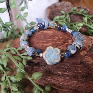 May include: A handcrafted bracelet featuring a variety of blue ceramic beads, including a large flower-shaped bead. The bracelet incorporates silver-toned metal accents and a small blue tassel. The beads vary in size and texture.
