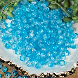 Pode incluir: Um conjunto de contas de vidro azul claro, com acabamento craquelado. As contas s&atilde;o redondas e de tamanhos variados, com um aspeto transl&uacute;cido. Est&atilde;o dispostas num prato decorativo azul claro, com folhagem verde no fundo. Ideal para a cria&ccedil;&atilde;o de joias ou projetos de artesanato.