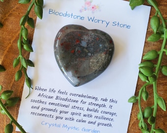 May include: A heart-shaped bloodstone worry stone with red and gray patterns. The stone rests on a white card with text that reads "Bloodstone Worry Stone" and a description of its properties. Green vines frame the scene.