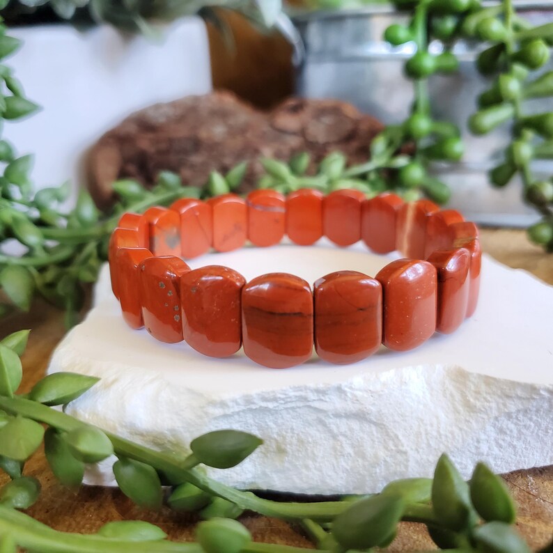 K&ouml;nnte beinhalten: Ein Armband aus rotem Jaspis mit rechteckigen Perlen. Das Armband wird auf einer wei&szlig;en, strukturierten Oberfl&auml;che pr&auml;sentiert, umgeben von gr&uuml;nem Laub. Die Perlen haben eine polierte Oberfl&auml;che und variieren von hell- bis dunkelrot.