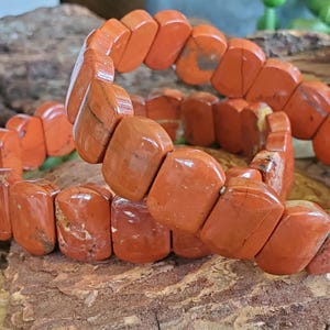 K&ouml;nnte beinhalten: Ein Armband aus orange-roten, rechteckigen Perlen. Die polierten Perlen sind zu einem flexiblen Band aufgereiht. Das Armband wird auf einer nat&uuml;rlichen Holzoberfl&auml;che pr&auml;sentiert. Die Perlen sind etwa 1,5 cm lang.