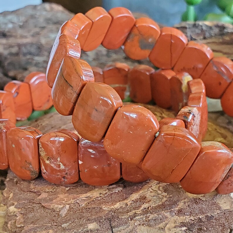 K&ouml;nnte beinhalten: Ein Armband aus orange-roten, rechteckigen Perlen. Die polierten Perlen sind zu einem flexiblen Band aufgereiht. Das Armband wird auf einer nat&uuml;rlichen Holzoberfl&auml;che pr&auml;sentiert. Die Perlen sind etwa 1,5 cm lang.