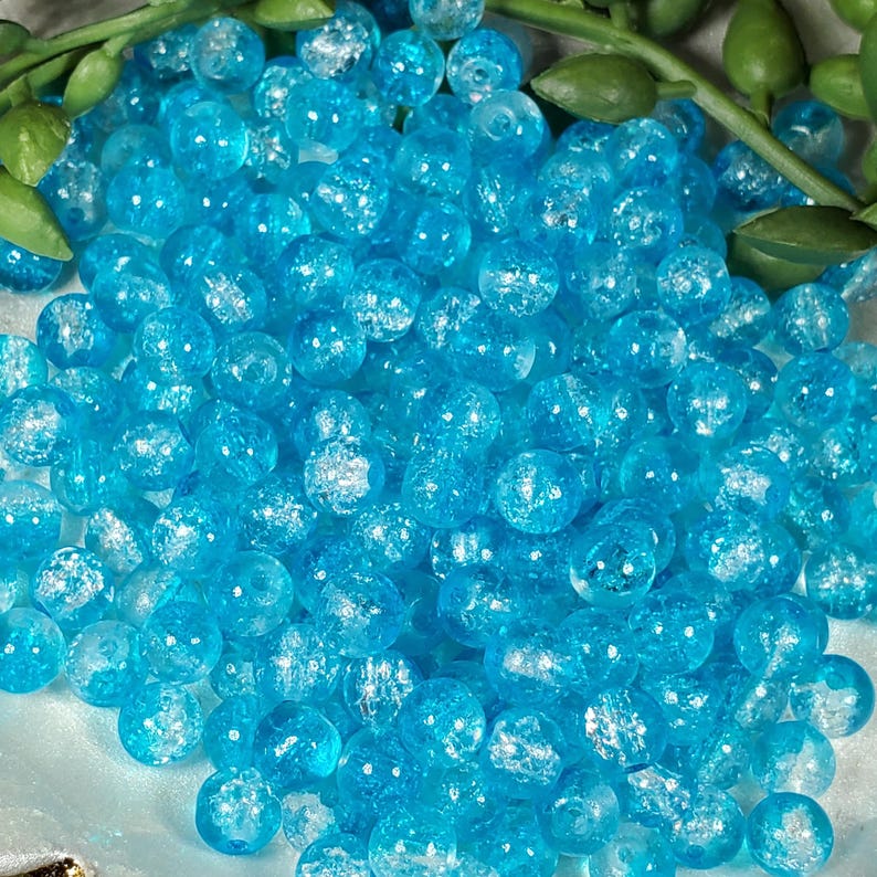 Pode incluir: Uma foto em close-up de uma pilha de contas de vidro azul claro com efeito craquelado. As contas s&atilde;o redondas e de tamanhos variados. Folhas verdes s&atilde;o vis&iacute;veis no fundo, adicionando um contraste natural &agrave;s contas azuis brilhantes. Estas contas s&atilde;o ideais para fazer joias.