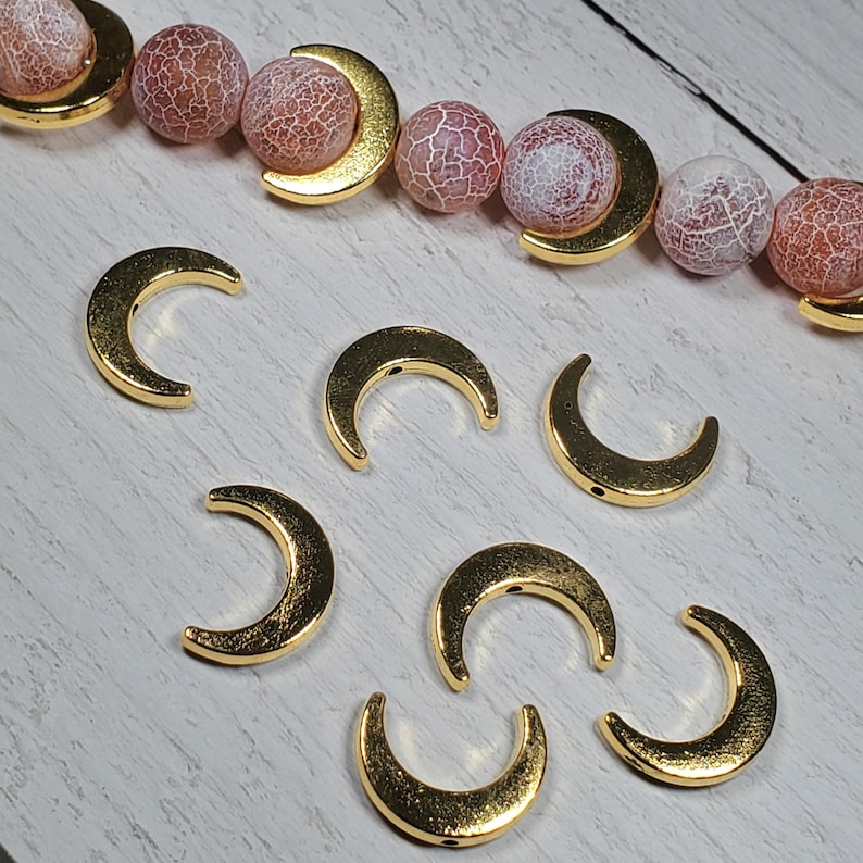 Puede incluir: Encantos dorados en forma de media luna y una hebra de cuentas con piedras estampadas en rosa y blanco. Los encantos est&aacute;n dispuestos sobre una superficie clara con vetas de madera. Las cuentas se combinan con acentos dorados de media luna.