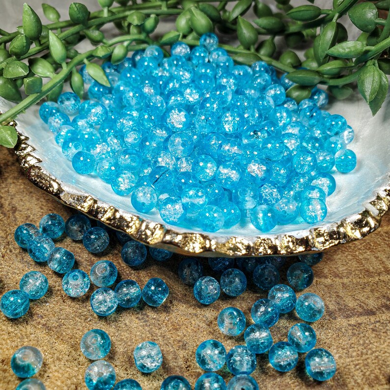 Pode incluir: Uma cole&ccedil;&atilde;o de contas de vidro azul brilhante com acabamento craquelado. As contas est&atilde;o espalhadas sobre uma superf&iacute;cie texturizada e numa concha decorativa com borda dourada. A folhagem verde &eacute; vis&iacute;vel no fundo.