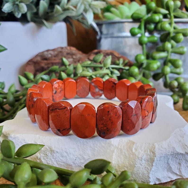 K&ouml;nnte beinhalten: Ein Armband aus rotem Jaspis mit facettiertem Design. Das Armband besteht aus oval geformten Perlen mit poliertem Finish. Die Perlen sind tiefrot mit dunkleren roten und schwarzen Einschl&uuml;ssen. Das Armband liegt auf einer wei&szlig;en, strukturierten Oberfl&auml;che.