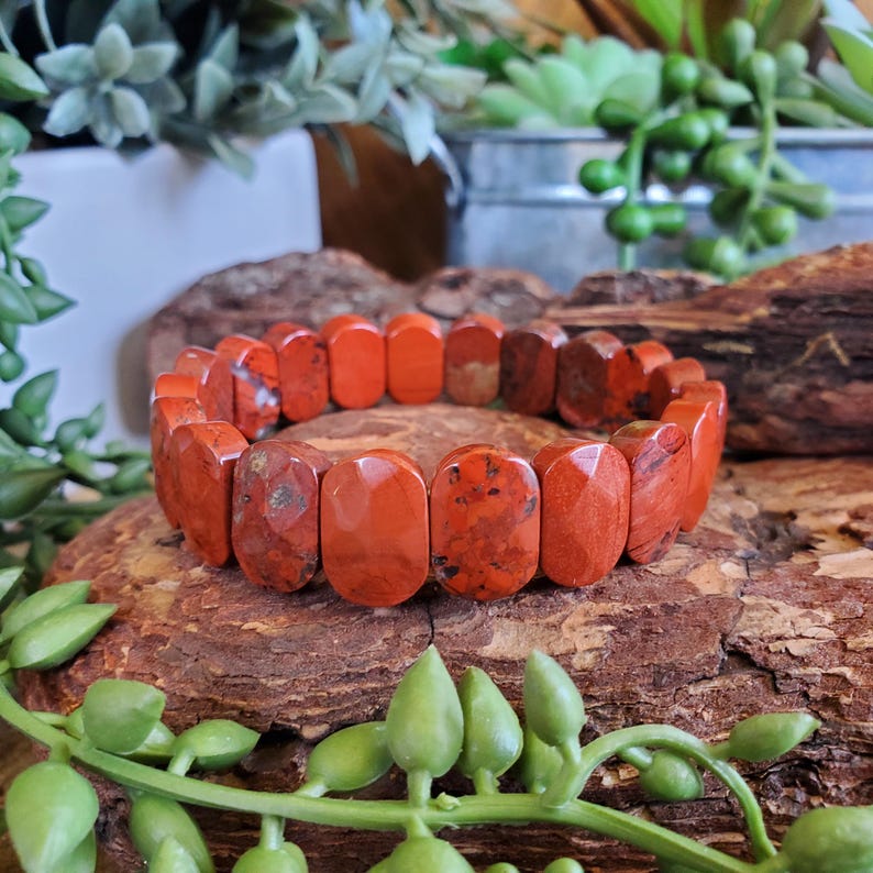 K&ouml;nnte beinhalten: Ein rotes Jaspis-Armband mit rechteckigen Perlen. Das Armband besteht aus polierten, rot-orangen Steinen. Die Perlen sind zu einem flexiblen Band aufgereiht. Das Armband wird auf einem Holzst&uuml;ck mit gr&uuml;nen Pflanzen im Hintergrund pr&auml;sentiert.