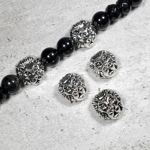 May include: A beaded bracelet with black onyx beads and silver lion head charms. The lion head charms are detailed with a textured mane and facial features. Additional loose lion head charms are scattered nearby.