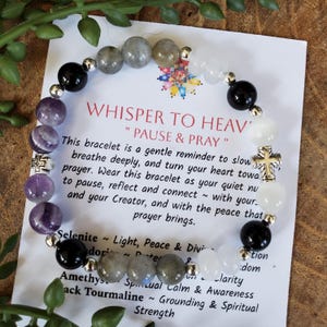 May include: A beaded bracelet with a mix of black, white, gray, and purple stones. The bracelet is next to a card with the text "WHISPER TO HEAVEN" and "PAUSE & PRAY". The card also lists the properties of the stones.