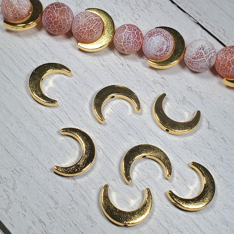 Puede incluir: Amuletos y cuentas de media luna doradas. Los amuletos est&aacute;n dispuestos sobre una superficie clara. Las cuentas son rosas y blancas con detalles de media luna dorados. Estos componentes son adecuados para la fabricaci&oacute;n de joyas y la artesan&iacute;a.