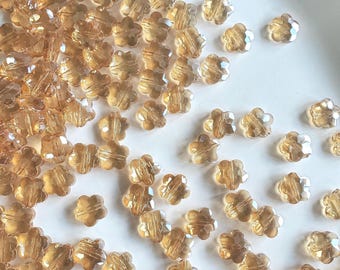 May include: A collection of small, translucent, light brown flower-shaped beads. Each bead features a faceted surface that reflects light, creating a sparkling appearance. The beads are scattered across a plain, light-colored surface.