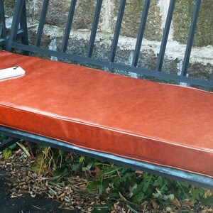 May include: A brown leather cushion sits on a black metal bench. The bench is made of wrought iron and has a simple design. The cushion is thick and comfortable, and the bench is perfect for relaxing outdoors.