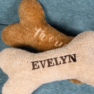 May include: Two plush dog bone shaped pillows, one brown with the word "fur-ever" embroidered on it and one beige with the name "EVELYN" embroidered on it.