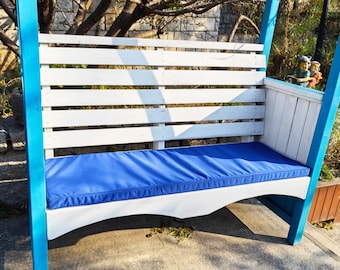 May include: A white wooden bench with blue painted supports and a blue cushion. The bench is attached to a wooden pergola.