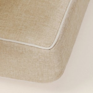 May include: Close-up of a beige fabric cushion with a white piping detail around the edge.