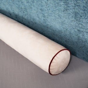 May include: A cream-colored cylindrical pillow with a red trim. The pillow is lying on a gray surface.