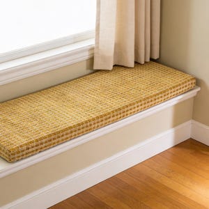 May include: A rectangular window seat cushion with a woven yellow and cream pattern. The cushion is placed on a white window ledge, with a beige curtain visible in the background. The floor is made of wood.