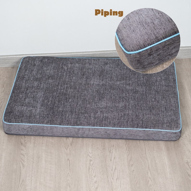 Pu&ograve; includere: Un letto per cani rettangolare grigio con una bordatura blu chiaro attorno ai bordi. Il letto &egrave; su un pavimento di legno. La parola "Piping" &egrave; scritta in testo marrone sopra il letto.