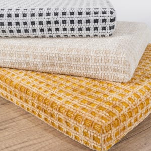 May include: Three textured fabric cushions stacked on top of each other. The top cushion is a light gray with a black square pattern. The middle cushion is a light beige. The bottom cushion is a golden yellow with a square pattern.