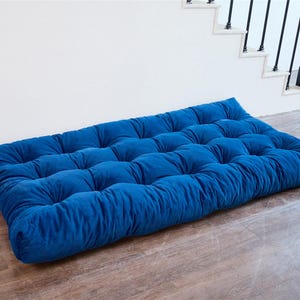 May include: A blue velvet floor cushion with tufted buttons. The cushion is rectangular and has a soft, plush texture.