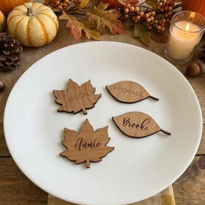 May include: Four laser-cut wooden leaf-shaped place cards resting on a white plate. Two are maple leaf shapes engraved with the names Jason and Annie, and two are oval leaf shapes engraved with the names Michael and Brooke in script lettering.
