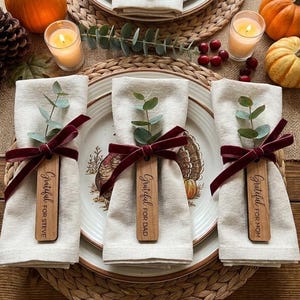 May include: Three cream-colored cloth napkins are each tied with a deep red velvet ribbon and a wooden tag. The tags are engraved with the text Grateful for Steve, Grateful for Dad, and Grateful for Mom, and each is topped with a sprig of green eucalyptus.