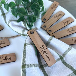 May include: Wooden name tags with names like "Marlowe", "Miles", and "Wendy" are arranged on a white plate with a green and white checkered napkin. The tags are light brown with a hole at the top and are perfect for a wedding or dinner party.