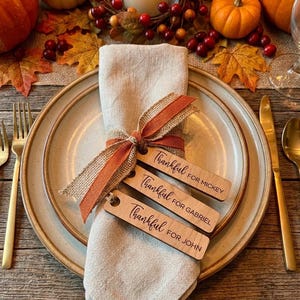 May include: A beige linen napkin folded on a ceramic plate, tied with a burlap and orange ribbon. Three rectangular wooden tags are attached, each engraved with the word Thankful followed by the names Mickey, Gabriel, and John respectively.