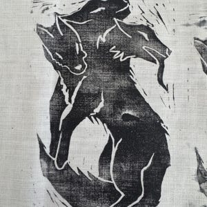 Peut inclure: Impression linogravée en noir et blanc sur tissu représentant trois loups stylisés. L'œuvre d'art représente les animaux dans une pose dynamique, avec un travail de ligne détaillé créant texture et forme. L'impression est sur fond blanc.