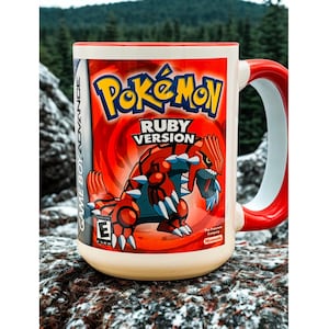 May include: A ceramic mug featuring the Pokémon Ruby Version game cover art. The mug has a red handle and rim, with a cream-colored body. The design includes the Pokémon logo and a depiction of Groudon, a red and blue creature.