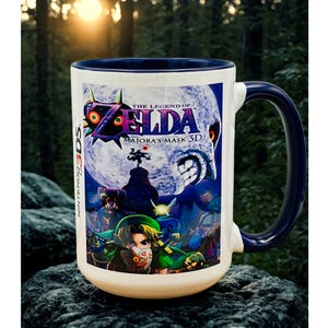 May include: White ceramic mug with a navy blue handle and rim. The mug features artwork from "The Legend of Zelda: Majora's Mask 3D" video game, including the game's logo and characters. The mug is set against a blurred forest background.