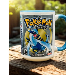 May include: White ceramic mug featuring the Pokémon Silver Version design. The mug has a blue rim and handle, with the Pokémon logo and a blue and silver Lugia graphic. The text "Gotta catch 'em all!" is also visible.