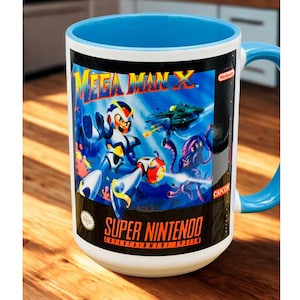 May include: White ceramic mug with a blue interior and handle, featuring a vibrant graphic from the Super Nintendo game Mega Man X. The design includes characters, a spaceship, and the game's logo. A perfect gift for gamers.