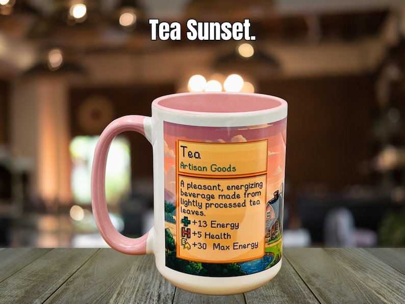 May include: A white ceramic mug with a pink handle and rim, featuring a label that reads "Tea Artisan Goods." The label describes the tea as an energizing beverage. The mug has the text "Tea Sunset" at the top.
