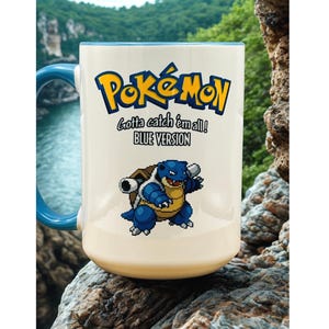 May include: A white ceramic mug with a blue handle and interior rim. The front features the yellow and blue Pok&eacute;mon logo, the text Gotta catch 'em all! BLUE VERSION, and a pixelated graphic of the blue turtle-like creature Blastoise.