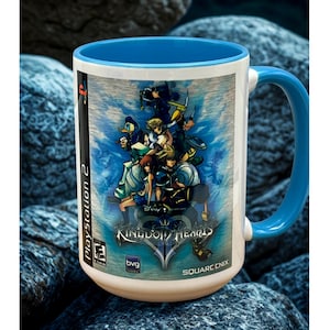 May include: White ceramic mug with a blue interior and handle. The mug features a colorful graphic from the video game Kingdom Hearts 2, with characters and the game's logo. The mug has the PlayStation 2 logo and the Square Enix logo.