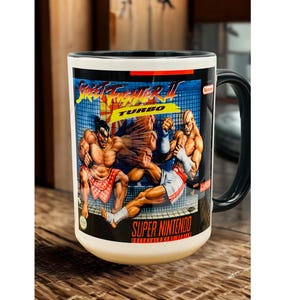 May include: A white ceramic mug with a black handle and interior, featuring a graphic print of the Street Fighter II Turbo video game cover art. The design displays two characters in a fighting stance with the text Street Fighter II Turbo.
