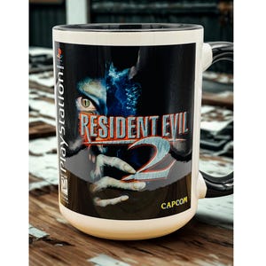 May include: A white ceramic mug featuring the Resident Evil 2 video game cover art. The mug has a black handle and rim, with the PlayStation logo on the side. The Capcom logo is also visible.