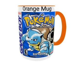 May include: A white ceramic mug with an orange handle and rim, featuring a graphic of the Pok&eacute;mon Blue Version Game Boy game cover. The design includes the Pok&eacute;mon logo, the text Gotta catch 'em all!, Blue Version, and Game Boy branding.