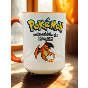 May include: A white ceramic mug with an orange handle and interior rim. The front features the blue and yellow Pok&eacute;mon logo, the text "Gotta catch 'em all!" and "RED VERSION", and a pixelated graphic of the orange dragon-like creature Charizard.