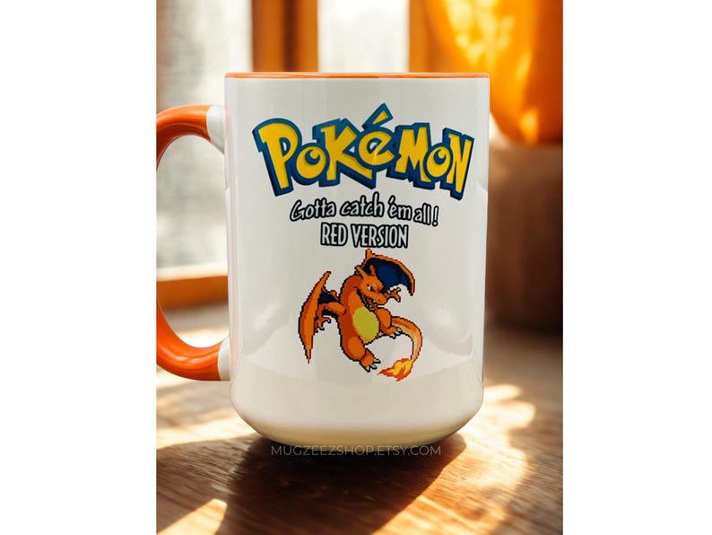 May include: A white ceramic mug with an orange handle and interior rim. The front features the blue and yellow Pok&eacute;mon logo, the text "Gotta catch 'em all!" and "RED VERSION", and a pixelated graphic of the orange dragon-like creature Charizard.