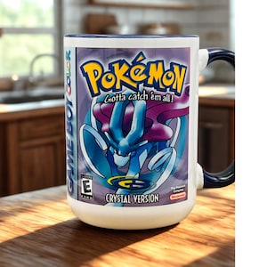 May include: A white ceramic mug with a dark blue handle and interior, featuring a graphic print of the Pokemon Crystal Version video game cover art, including the Pokemon Suicune, the Game Boy Color logo, and the text Gotta catch em all.