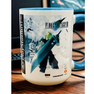 Op de afbeelding: Witte keramische mok met een blauwe binnenkant en handvat, met een Final Fantasy VII-ontwerp. De mok toont een personage met een groot zwaard, het logo van de game en het PlayStation-logo. De mok heeft een diameter van ongeveer 7,6 cm.