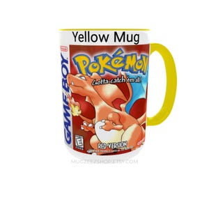 May include: A yellow ceramic mug featuring a graphic print of the Pok&eacute;mon Red Version Game Boy game cover. The design includes the orange Charizard character, the Pok&eacute;mon logo, the text Gotta catch 'em all!, and the Game Boy branding.