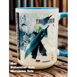 Op de afbeelding: Een witte keramische Final Fantasy VII-mok met een blauw handvat en rand. De mok heeft een karakterillustratie met een groot zwaard en het logo van de game. De tekst "PlayStation" is zichtbaar aan de zijkant. De mok is ongeveer 444 ml en is vaatwasser- en magnetronbestendig.