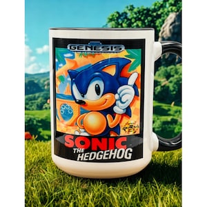 May include: A white ceramic mug with a black handle and interior, featuring a graphic of Sonic the Hedgehog on the front. The design includes the Sega Genesis logo at the top and the text Sonic The Hedgehog in red and white lettering below the character.