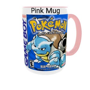May include: A pink ceramic mug featuring a graphic print of the Game Boy Pok&eacute;mon Blue Version video game cover art. The design includes the Pok&eacute;mon Blastoise, the Pok&eacute;mon logo, the text Gotta catch 'em all!, Blue Version, and an ESRB E rating.